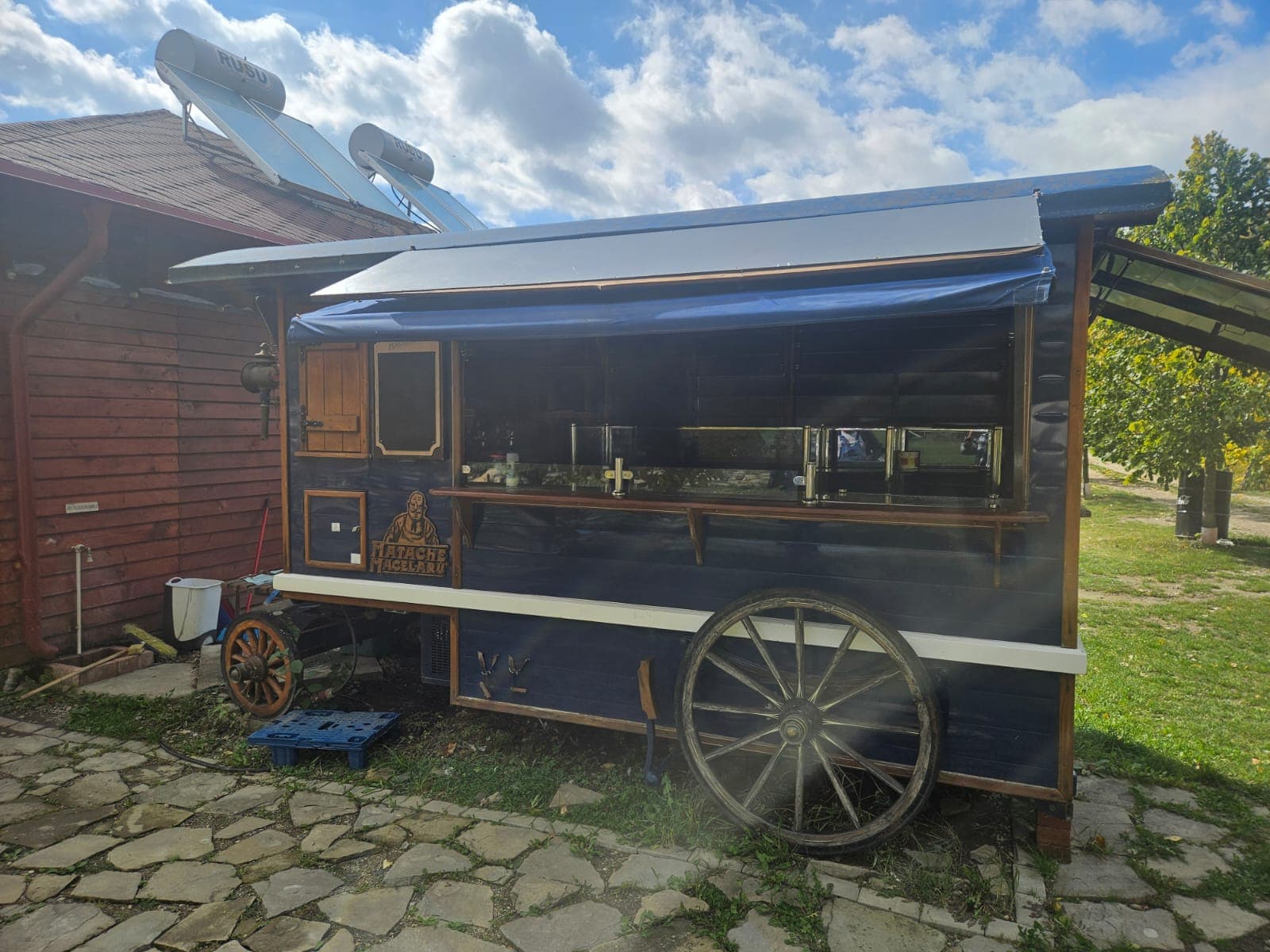 Blue rustic food trailer with large wooden wagon wheels parked on a stone path outdoors.