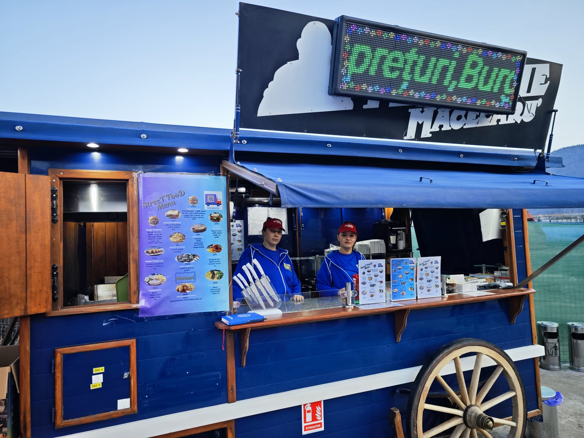 Two women in blue uniforms stand inside a blue food truck with a wooden wheel.
