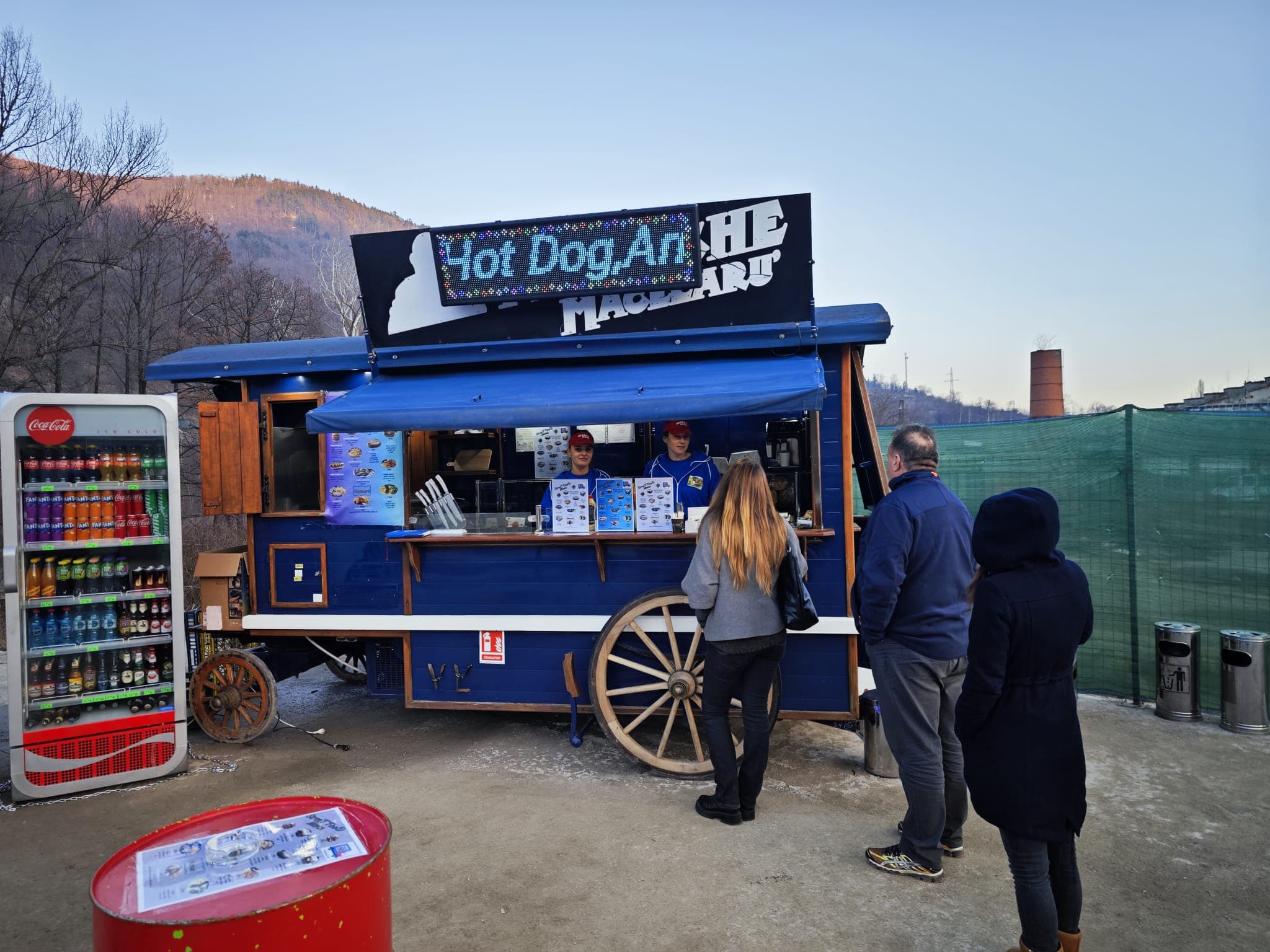 Customers queue at a blue wagon-style food truck with a 'Hot Dog Art' sign.