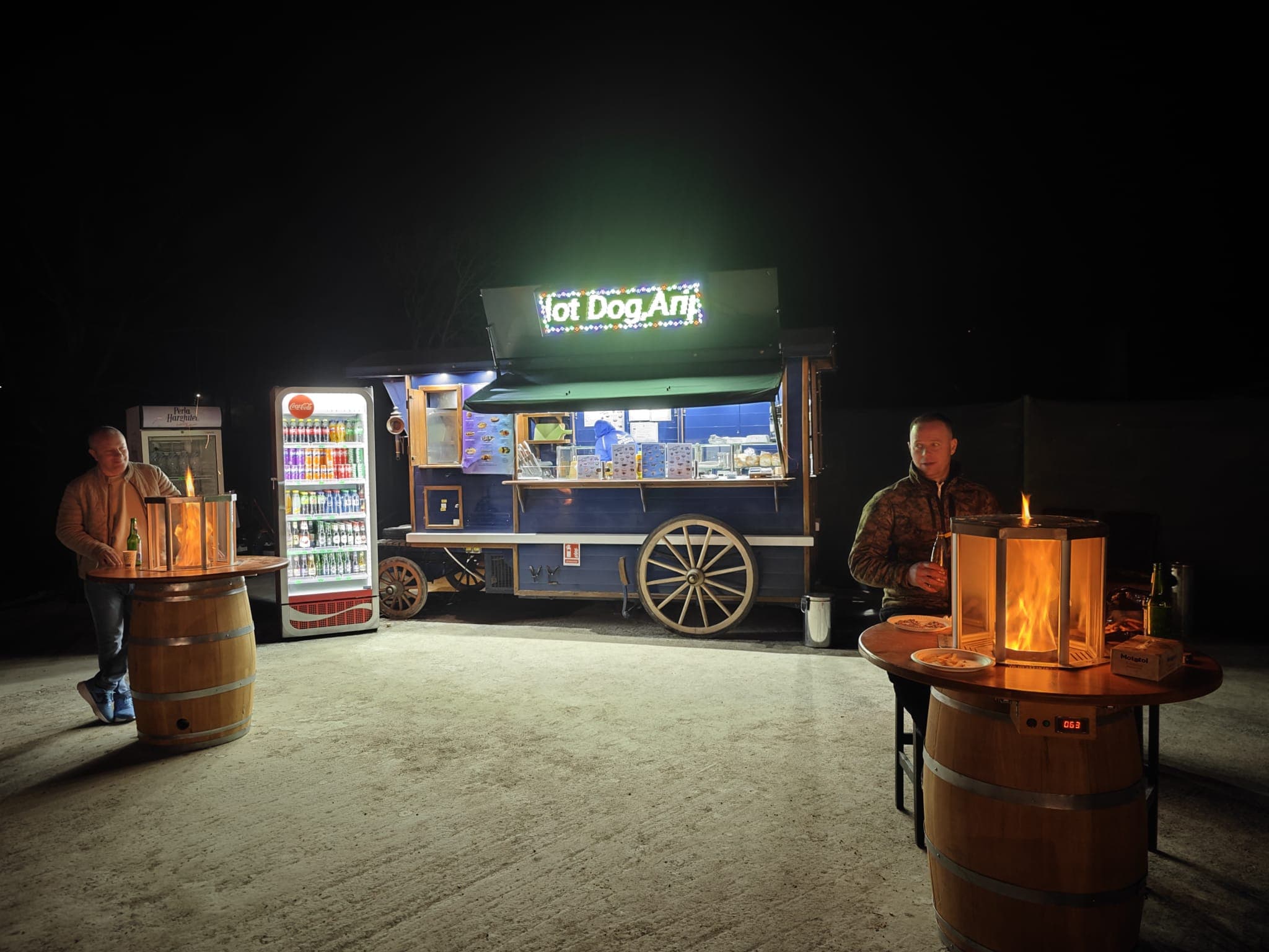 Blue hot dog truck at night with men standing at barrel tables with fire pits.