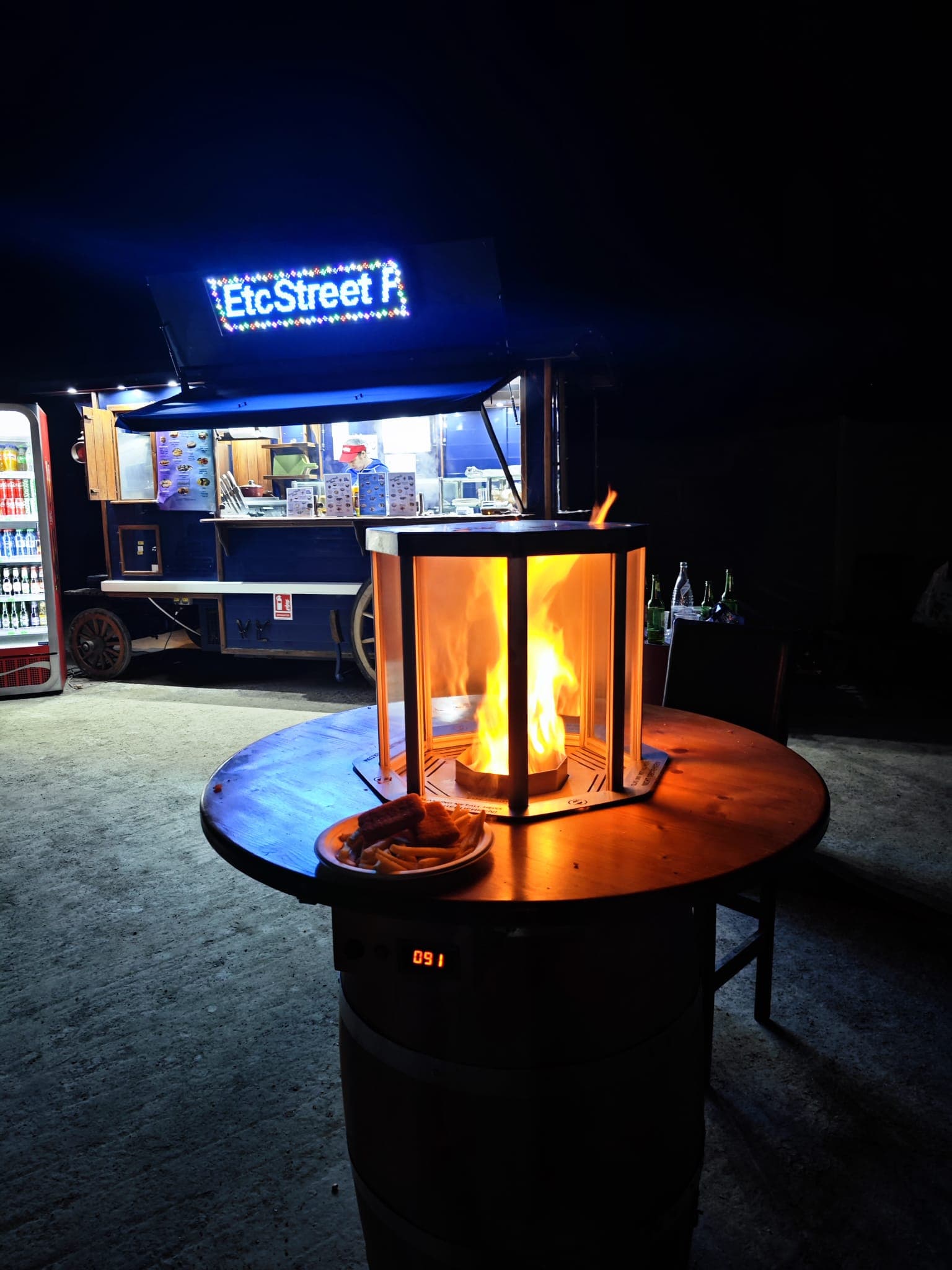 Barrel table with a glass fire pit and food, outside a food truck at night.