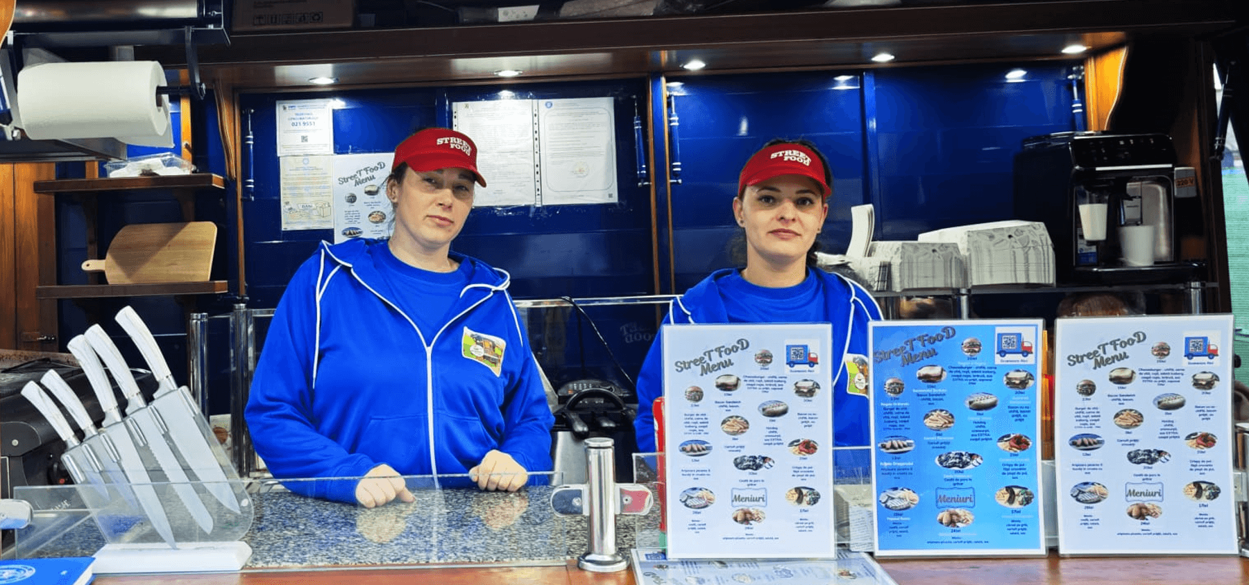 Two women in blue hoodies and red caps behind a street food counter with menus.